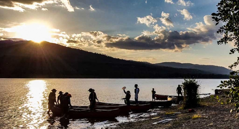 a group of outward bound students stand in shallow water beside their canoes as the sun is setting